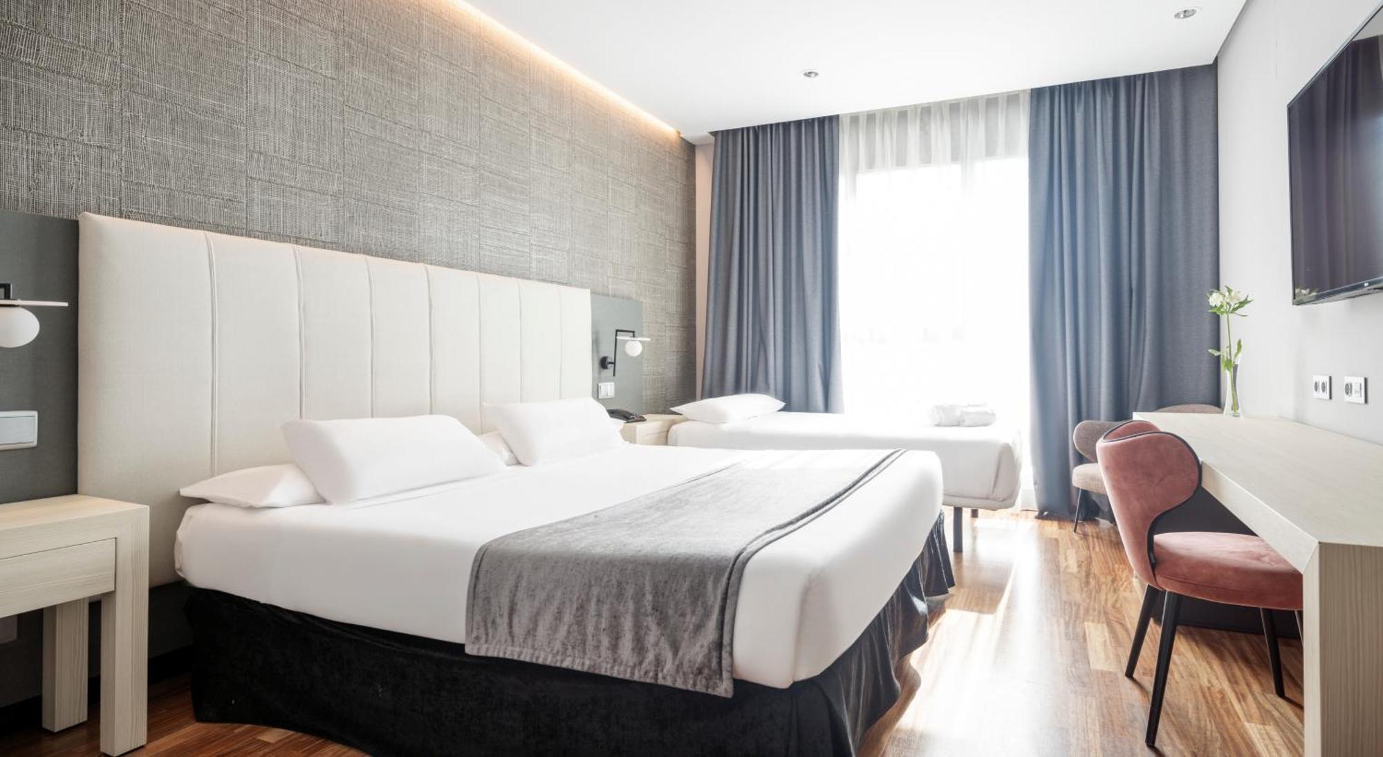 Ilunion Alcalá Norte Madri Quarto foto The photo shows a hotel room featuring two beds, one of which is a queen-sized bed with white linens and a gray throw blanket. Next to it, there is a smaller bed, possibly a twin. The room has a modern, minimalistic design with light wood flooring an